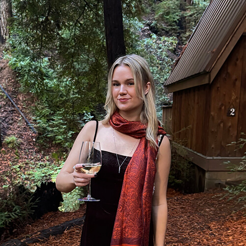 Blonde woman in black dress holding wine glass in a forest.